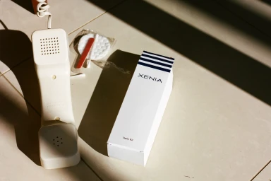 White landline telephone handset next to a branded Xenia vanity kit box and cotton pads on a tiled floor.
