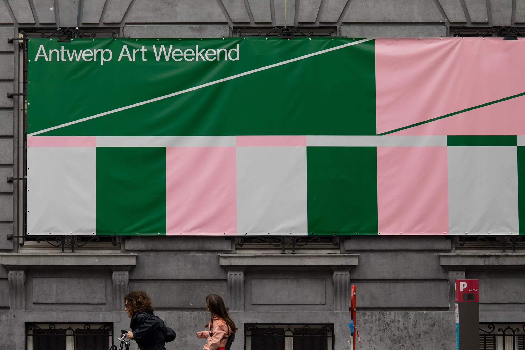 Large banner for Antwerp Art Weekend on building with pink, white, and green color blocks, two people walk by on the street below.