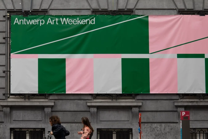 Large banner for Antwerp Art Weekend on building with pink, white, and green color blocks, two people walk by on the street below.