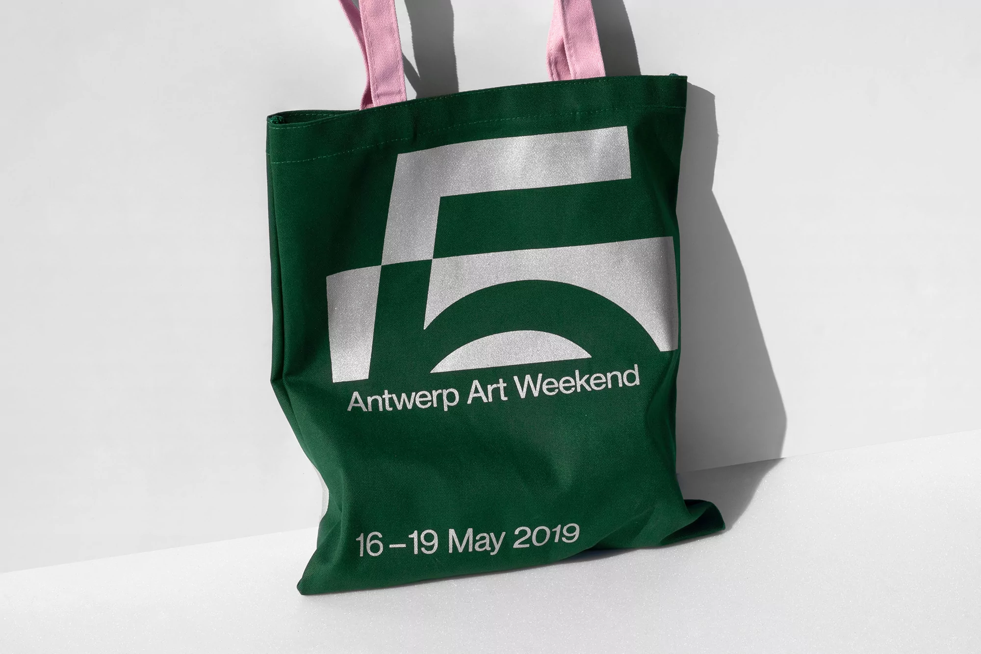 Green tote bag with 'Antwerp Art Weekend 16-19 May 2019' print, featuring pink handles, against a plain white background.