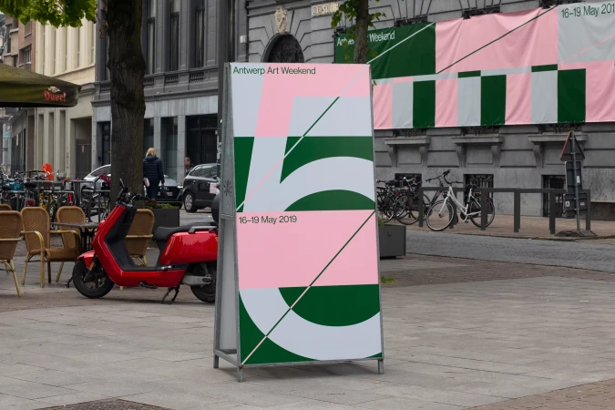 Art weekend advertisement on the city street with scooter, outdoor cafe, buildings and bikes in the background during the daytime.