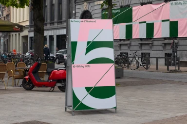 A promotional stand for Antwerp Art Weekend featuring graphic design in pink, green, and white on a city street.