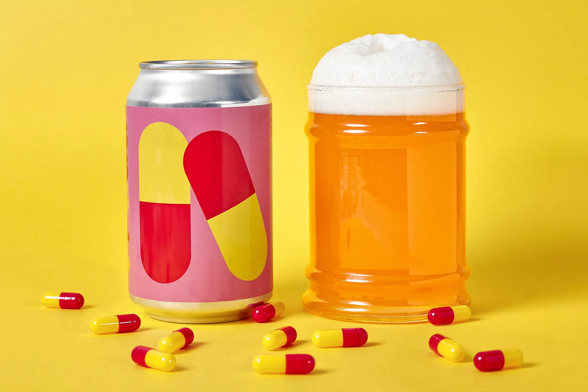 Stylized shot of a canned drink with a red and yellow pill design next to a mug of beer against a bright yellow backdrop.