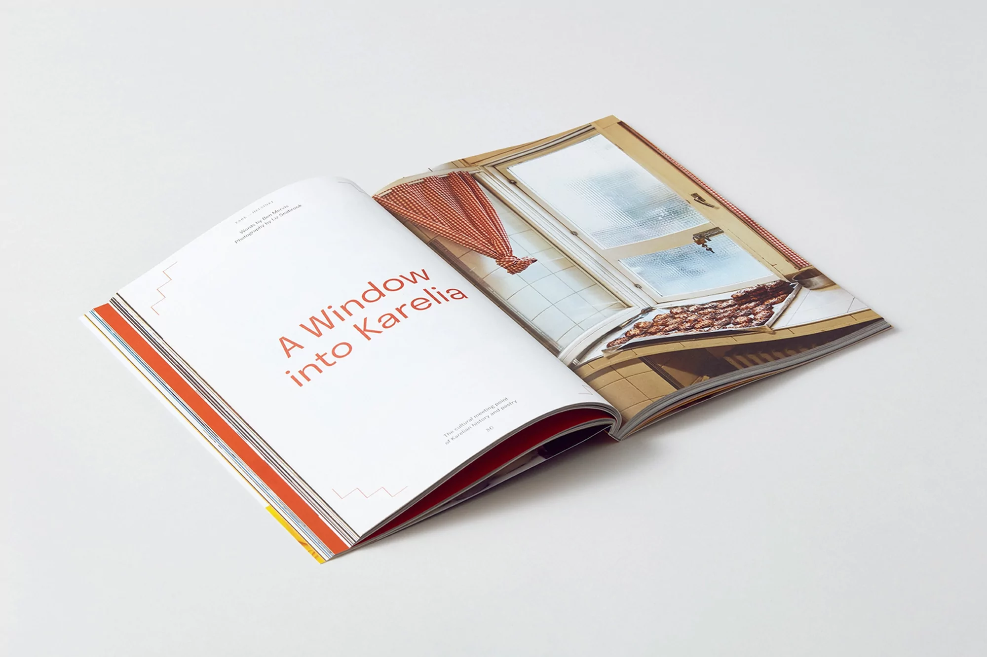 Open magazine spread features an interior shot of a window with red and white checkered curtains and a tray of baked goods.