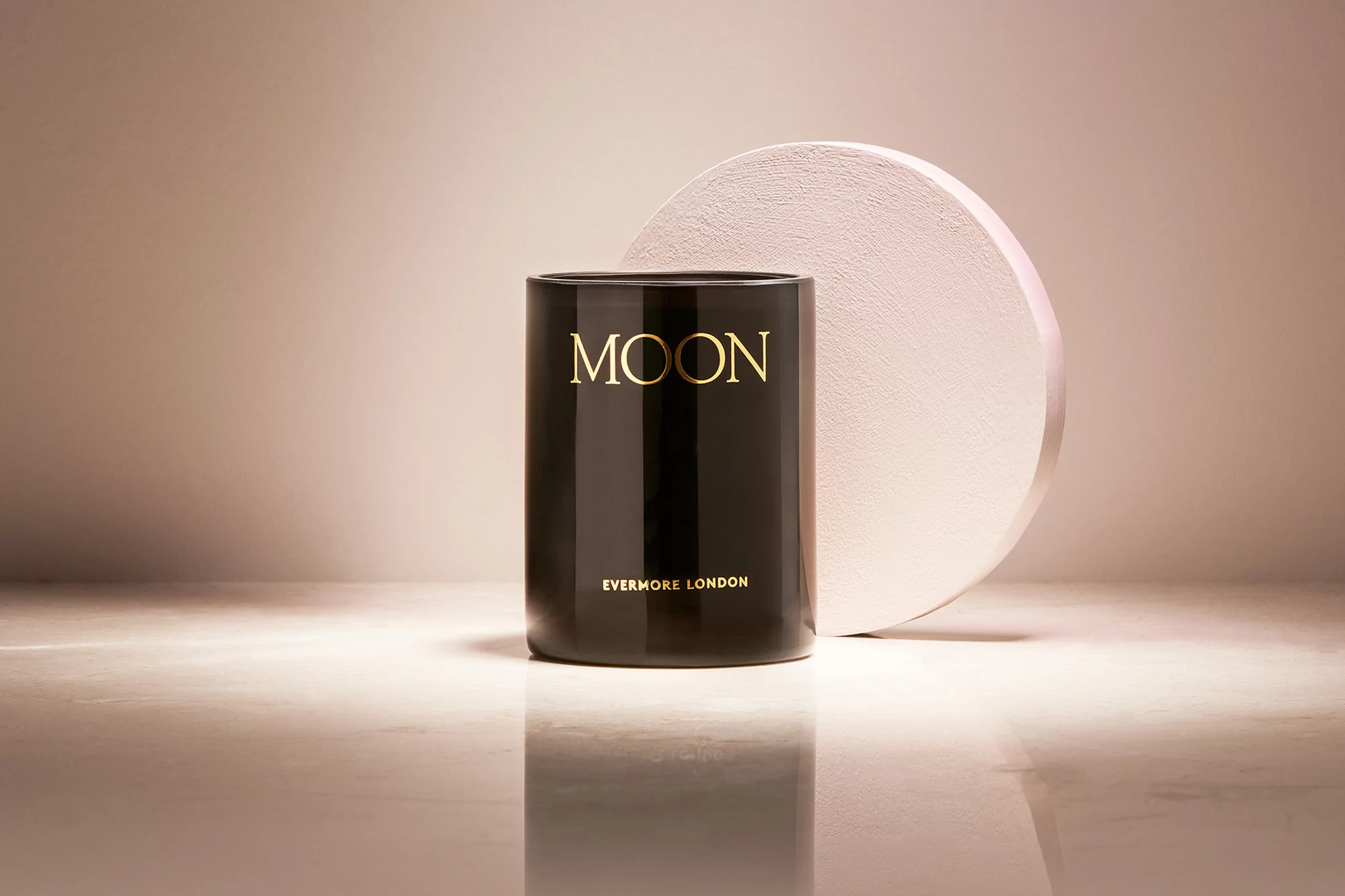 Minimalist product shot of a black candle with gold lettering reading 'Moon' next to a round, textured prop on a reflective surface.