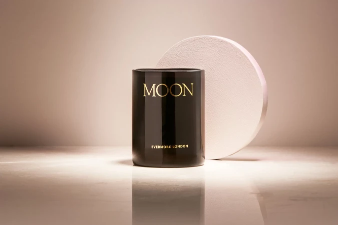Minimalist product shot of a black candle with gold lettering reading 'Moon' next to a round, textured prop on a reflective surface.