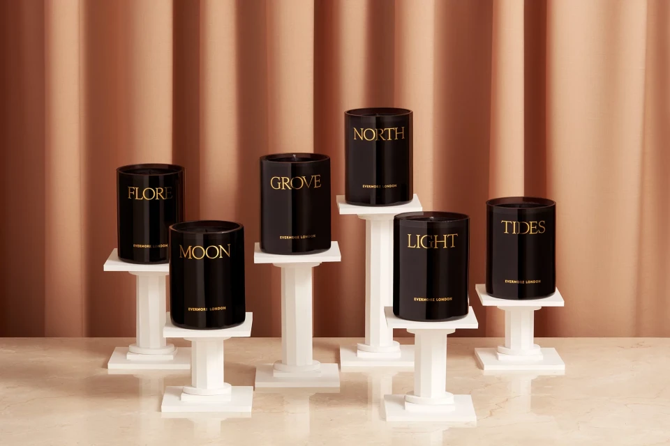 Several black Evermore London candles are displayed on white pedestals against a peach-colored draped fabric background in a product shot.