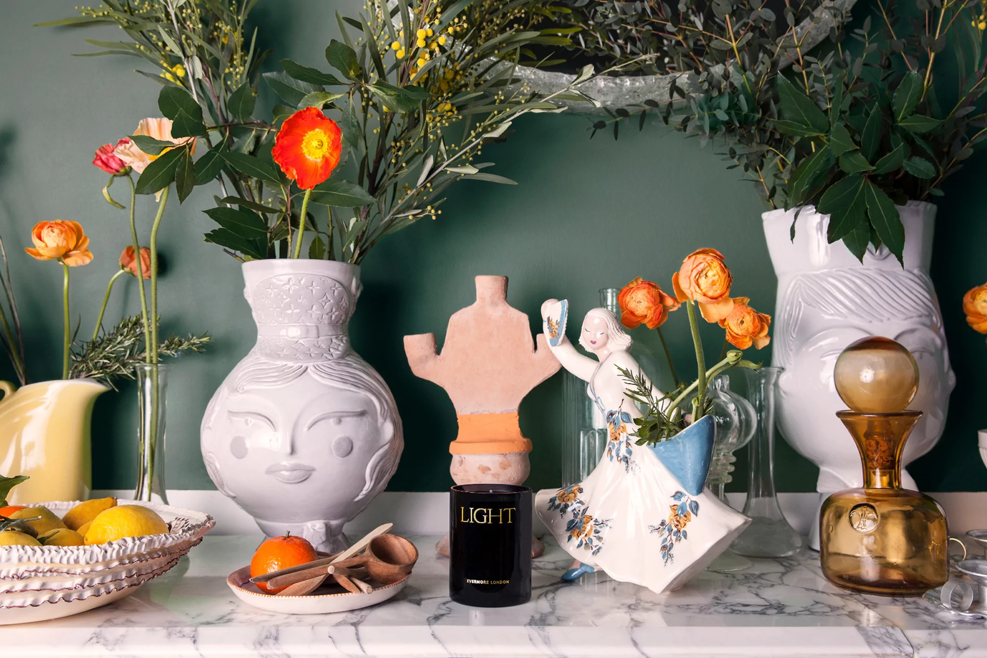 Still life featuring flowers in face-shaped vases, a candle, fruit, and a figurine in front of an emerald green wall.