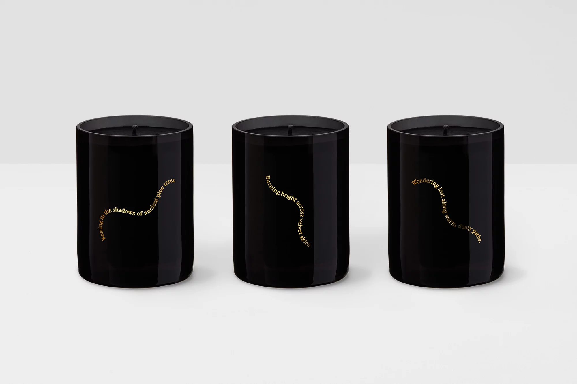 Three black tumbler candles with gold text labels, arranged side by side against a light background. Minimalist home decor.