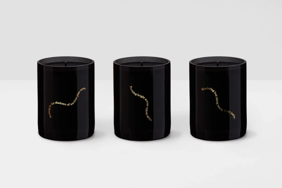 Three black tumbler candles with gold text labels, arranged side by side against a light background. Minimalist home decor.