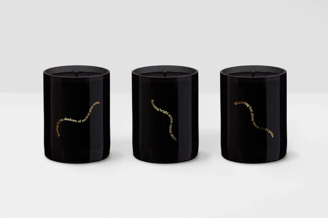 Three black tumbler candles with gold text labels, arranged side by side against a light background. Minimalist home decor.