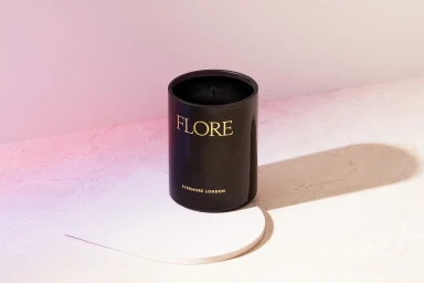 Black cylindrical candle jar with gold lettering on a geometric white platform against a pink-lit background.