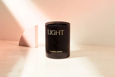 Black glass candle jar featuring gold text resting on a light marble surface with a diagonal beam of light.