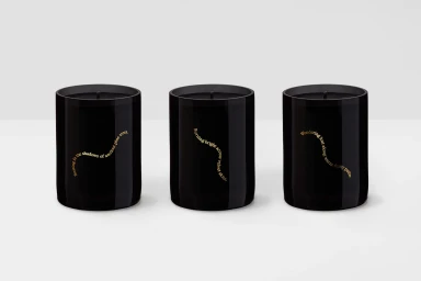 Three black glass candles with gold typography arranged in a row against a clean white seamless studio background.