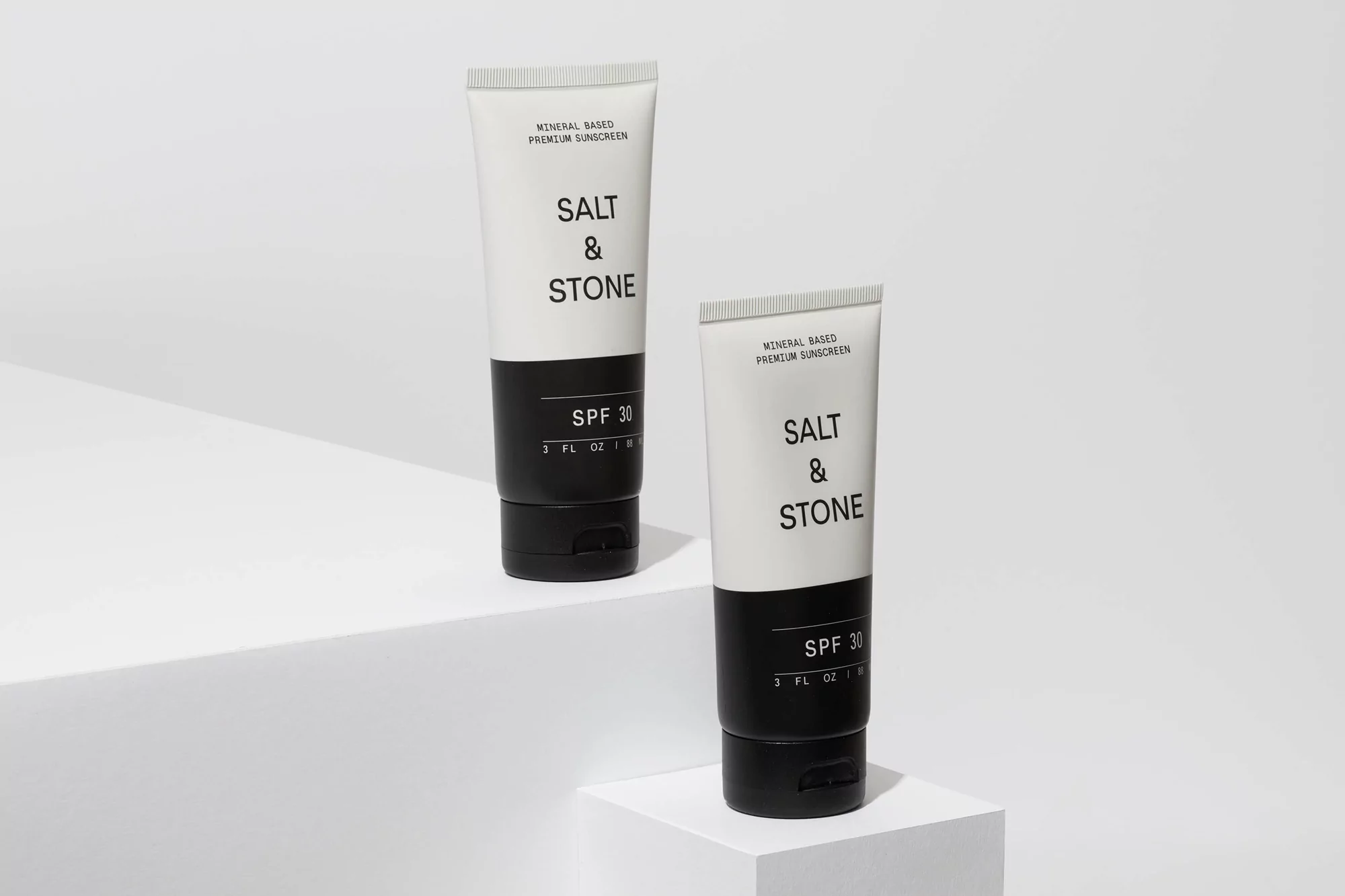 Two white tubes of Salt & Stone SPF 30 sunscreen sit on white geometric platforms against a white backdrop in a studio setting.