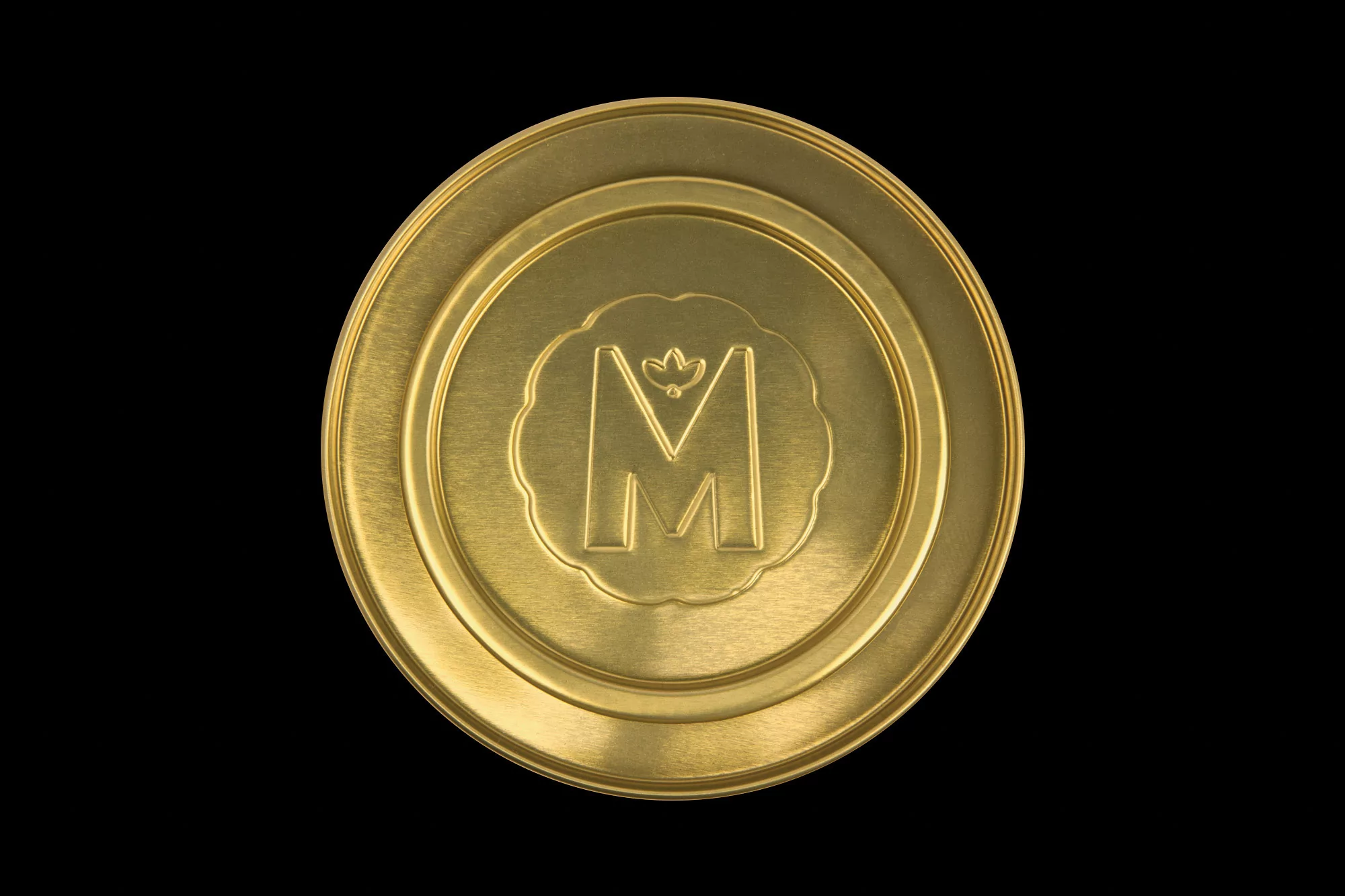Close-up studio shot of a golden disc with a central letter 'M' emblem on a black background, highlighting its metallic texture.