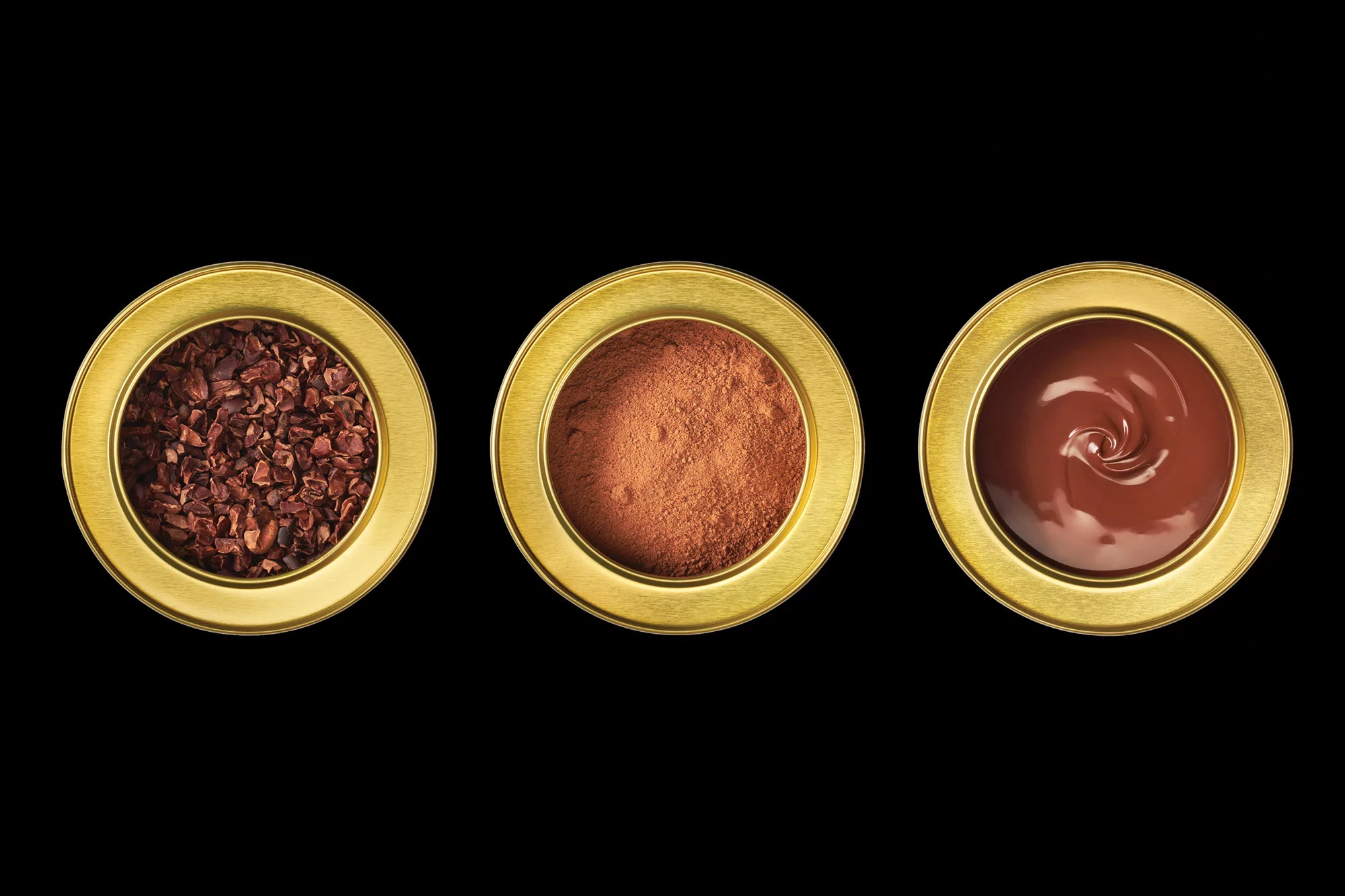 Close-up studio shot of three golden cups containing cacao nibs, cacao powder, and melted chocolate, set against a black background.