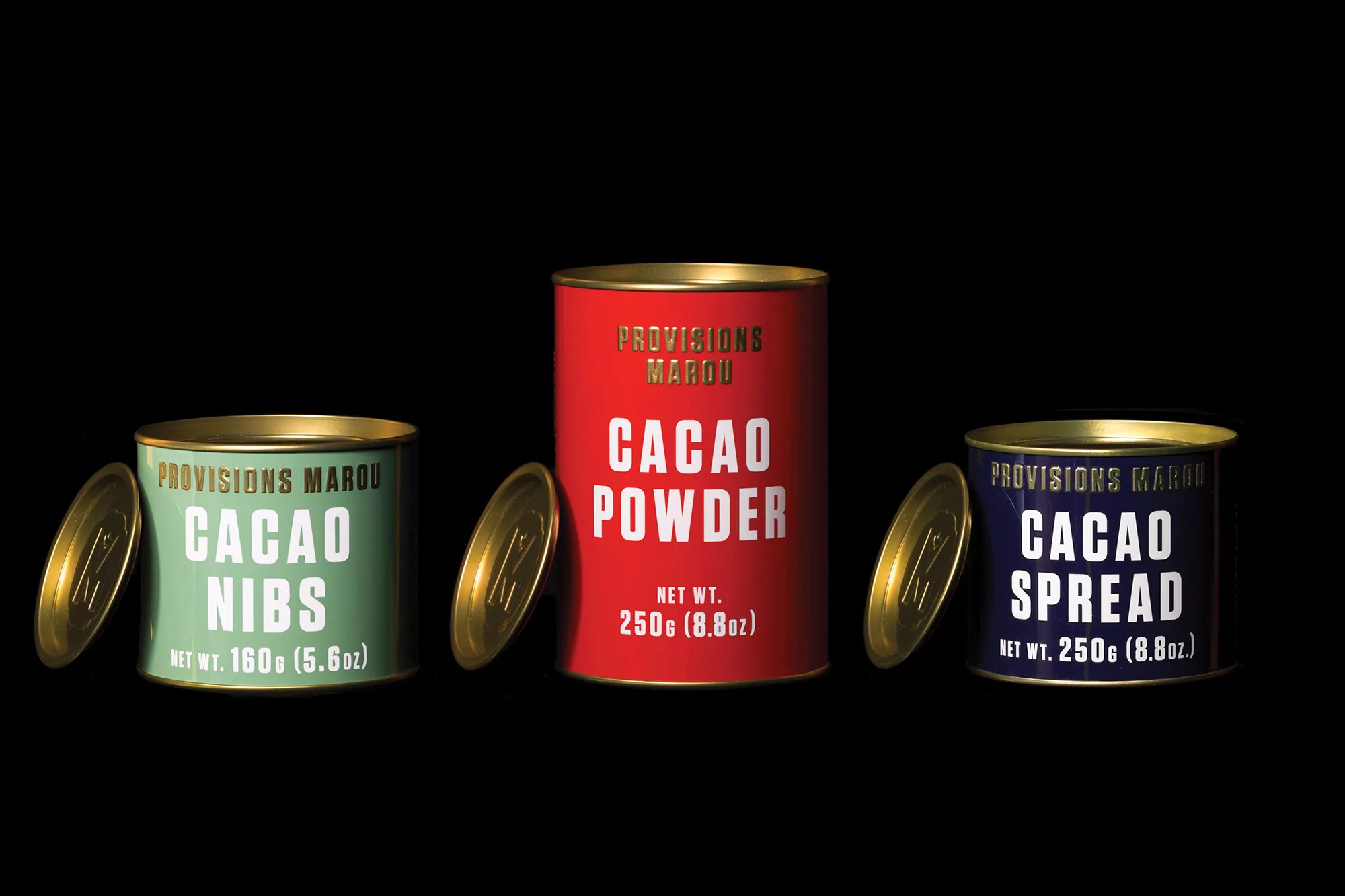 Three colorful Provisions Marou cacao cans against a black background: cacao nibs, cacao powder, and cacao spread.