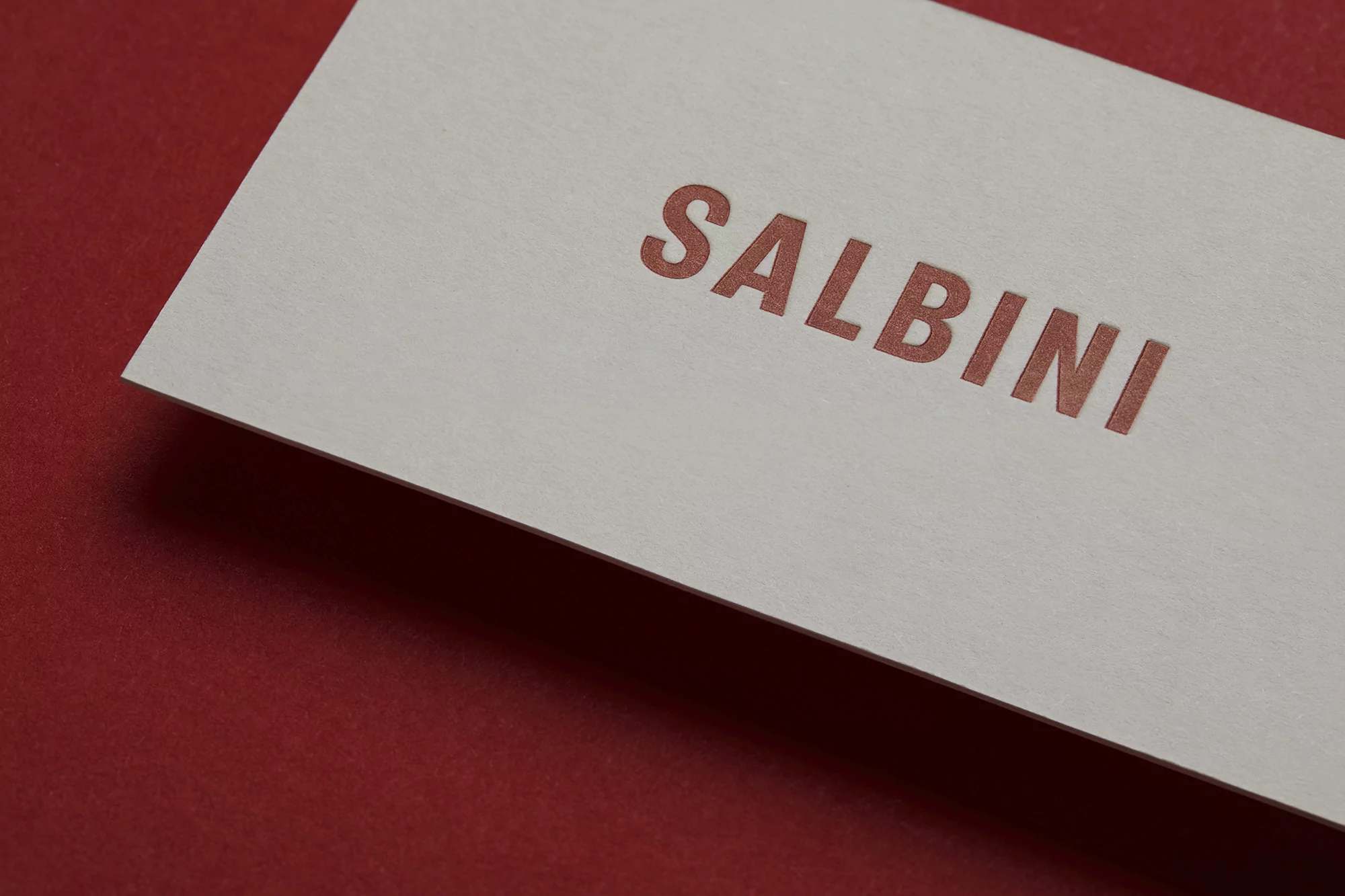 Close-up studio shot of a textured white card with the word 'SALBINI' in salmon letters, set against a vibrant red background.
