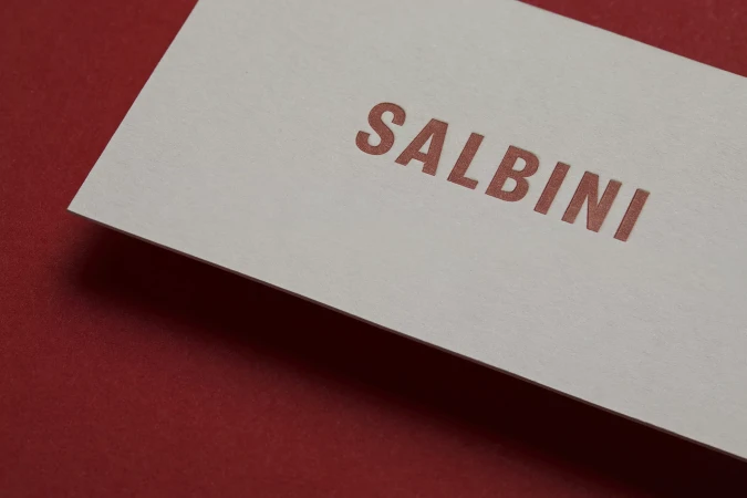 Close-up studio shot of a textured white card with the word 'SALBINI' in salmon letters, set against a vibrant red background.