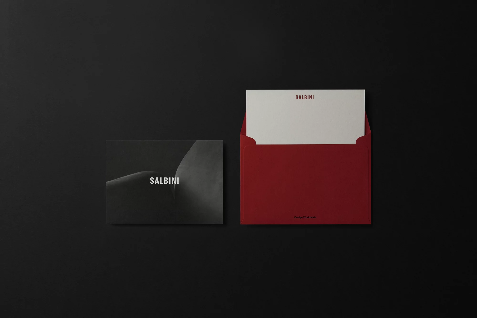 High-angle shot of a dark grey and black brochure with 'SALBINI' text, alongside a red envelope on a black backdrop.