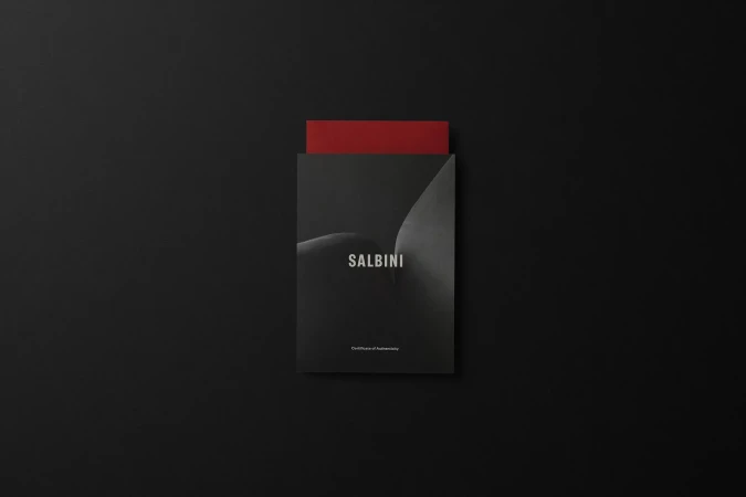 Flat lay of a black book with 'SALBINI' printed on the cover, partially revealing a red sheet of paper against a solid black background.
