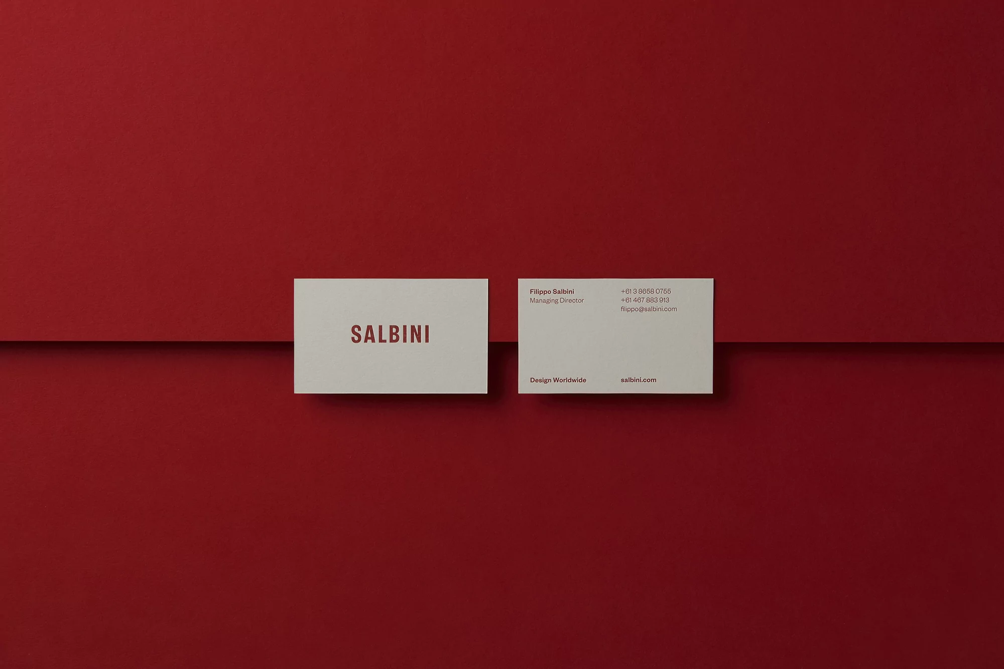 Two white SALBINI business cards on a red background with a horizontal line running behind them. Modern, minimalist design.