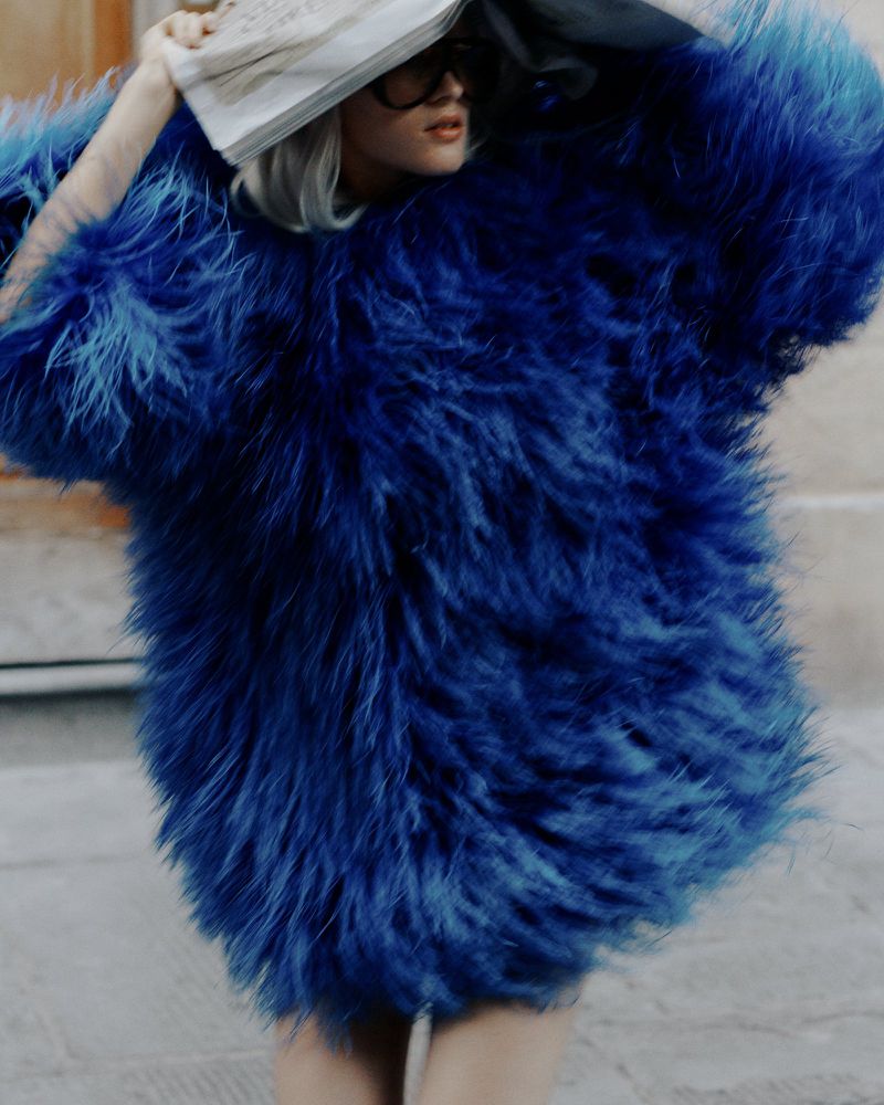 A woman in a large, bright blue faux fur coat stands outdoors with a newspaper held over her head, styled with dark glasses.