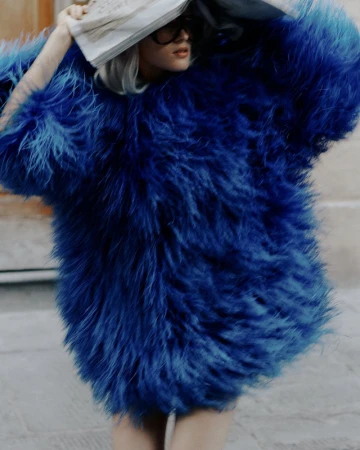 A woman in a large, bright blue faux fur coat stands outdoors with a newspaper held over her head, styled with dark glasses.
