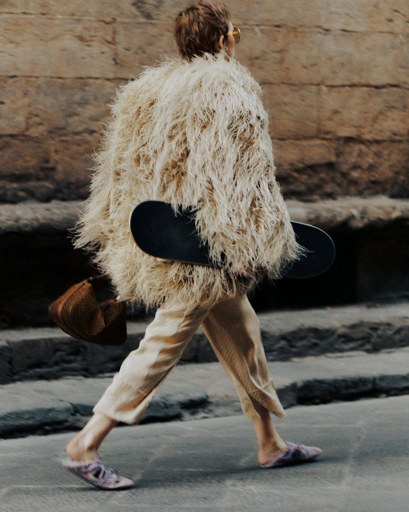 A person walks on a city street wearing a shaggy coat and carrying a skateboard, with tan pants and slipper shoes.