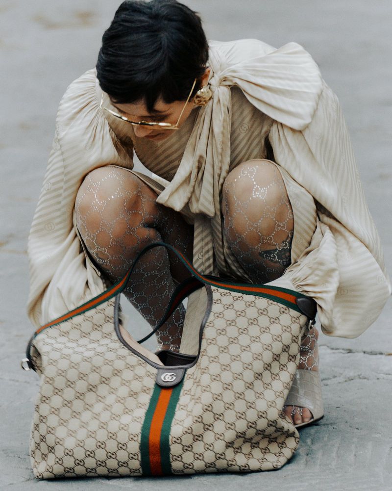 Stylish woman in patterned tights and a ruffled top crouches near a Gucci handbag, showcasing a blend of high fashion and accessory detail.