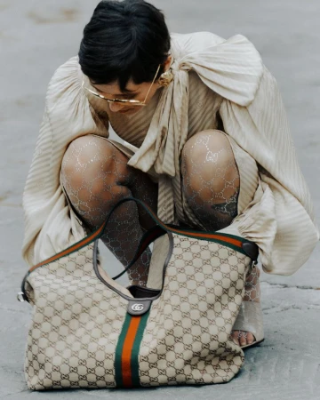 Stylish woman in patterned tights and a ruffled top crouches near a Gucci handbag, showcasing a blend of high fashion and accessory detail.