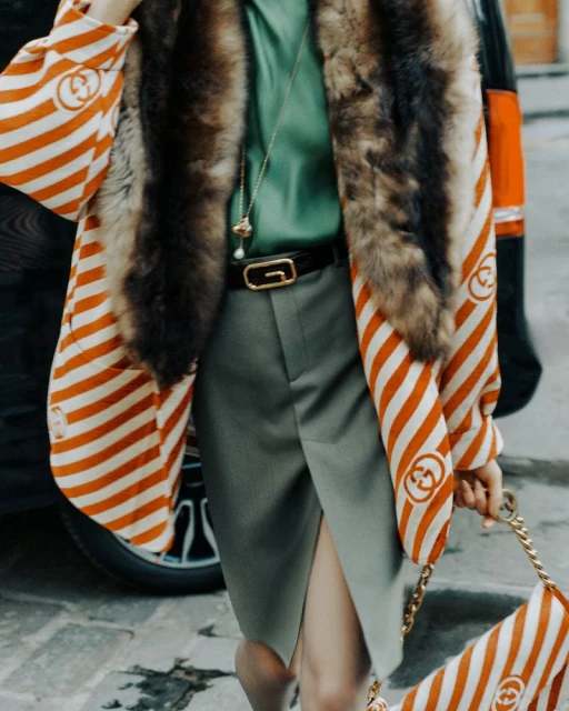 Woman in fur-trimmed coat, green top, and grey skirt carrying a striped bag, outdoors.