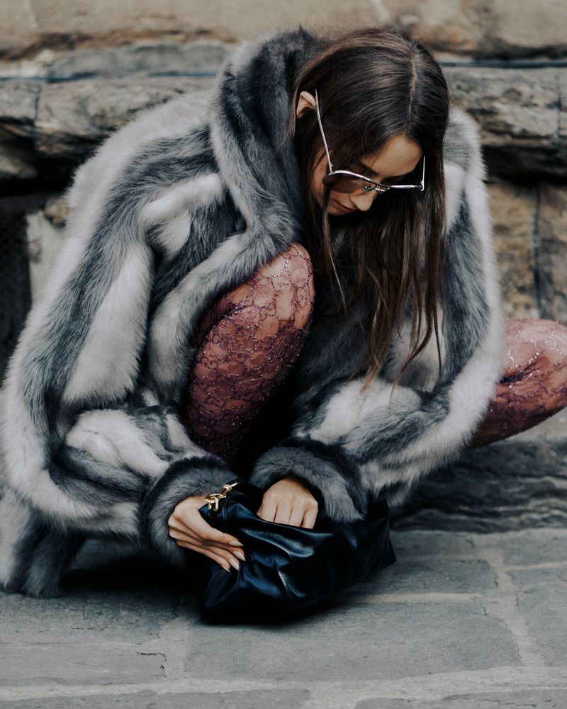Woman wearing fur coat and sunglasses squats with black purse on stone ground, posing in an outdoor fashion editorial setting.