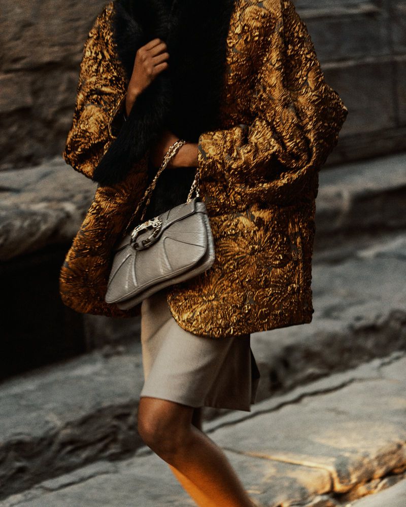 Woman in gold brocade jacket, fur collar, carrying gray purse on stone steps. Fashion editorial. Lifestyle photograph. Neutral colors.