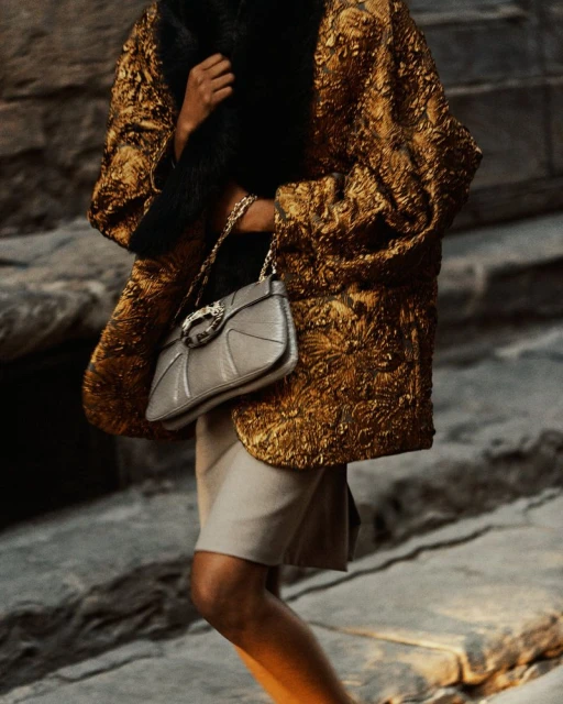 Woman in gold brocade jacket, fur collar, carrying gray purse on stone steps. Fashion editorial. Lifestyle photograph. Neutral colors.