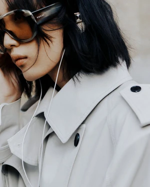 Close-up portrait of a woman wearing oversized sunglasses, wired earbuds, and a light-colored trench coat for Gucci Cruise 26.