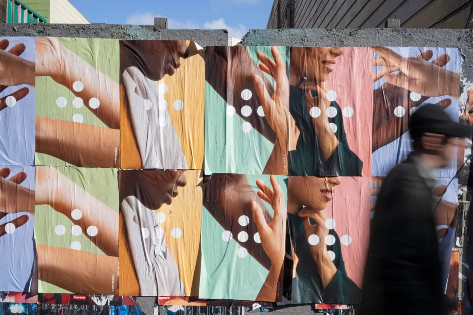 Street art installation of posters showing human arms, hands, and faces covered with white dots, with a blurred person walking past.