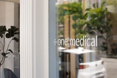 Storefront glass window displaying the One Medical brand logo with an interior view and greenery outside