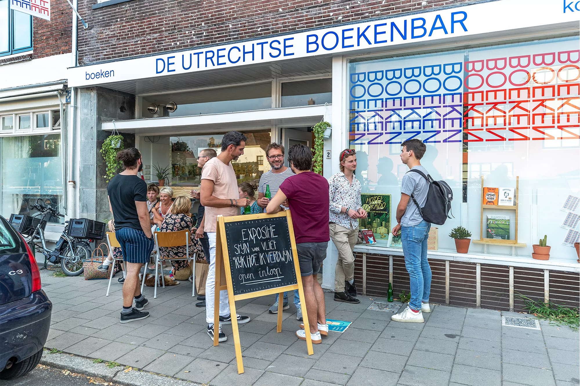Outdoor shot of a group of people standing in front of De Utrechtse Boekenbar bookstore with large windows and text on the storefront.