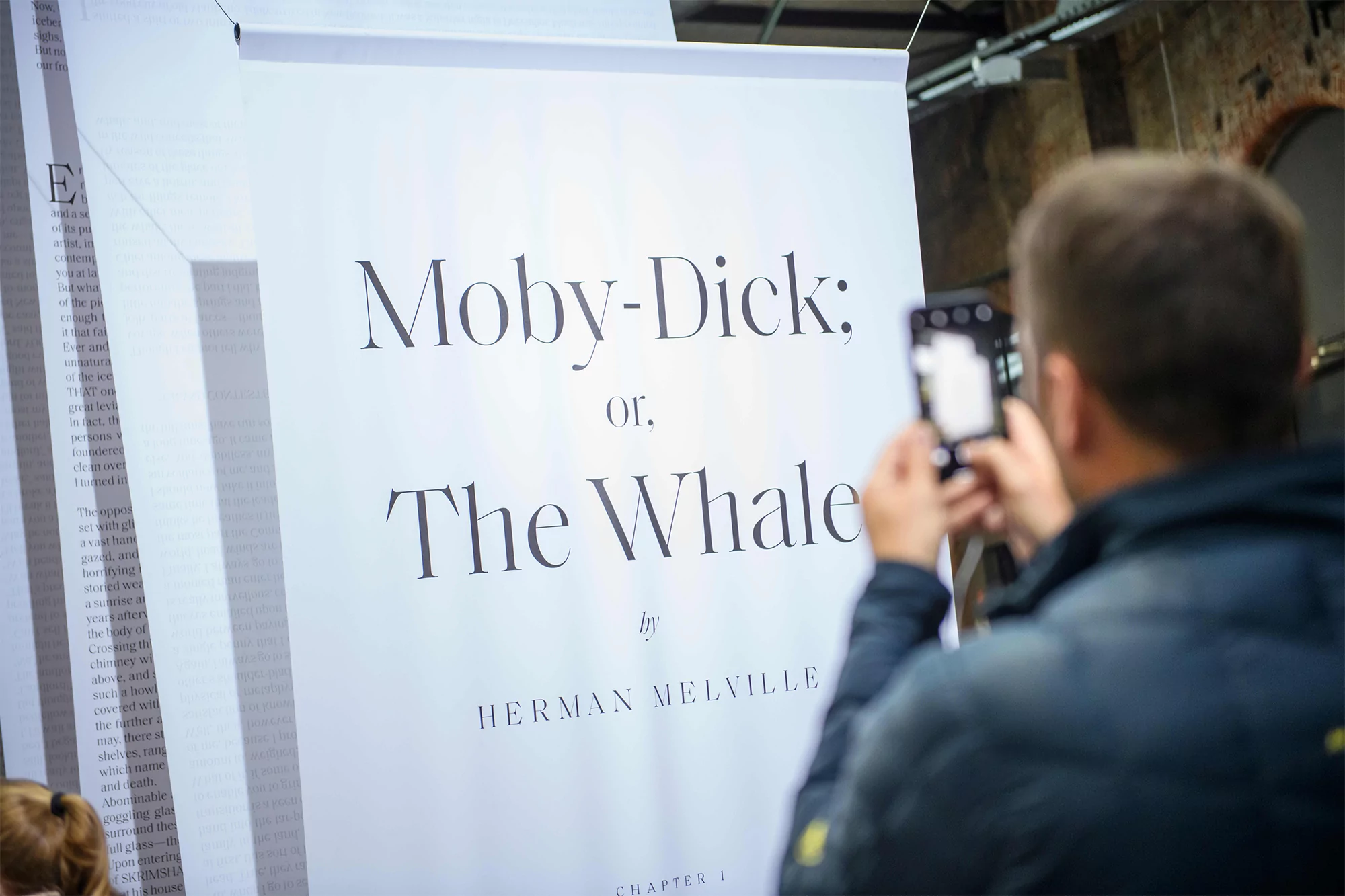 Man photographing a book poster titled "Moby-Dick; or, The Whale" by Herman Melville with a smartphone indoors.