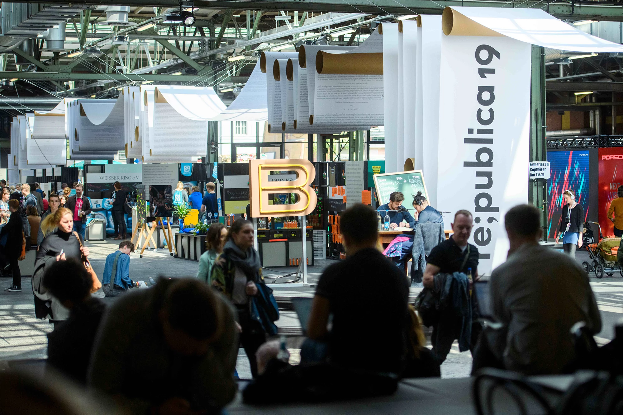 Large indoor convention hall with many attendees walking near vendor booths under white hanging paper banners; neutral color palette.