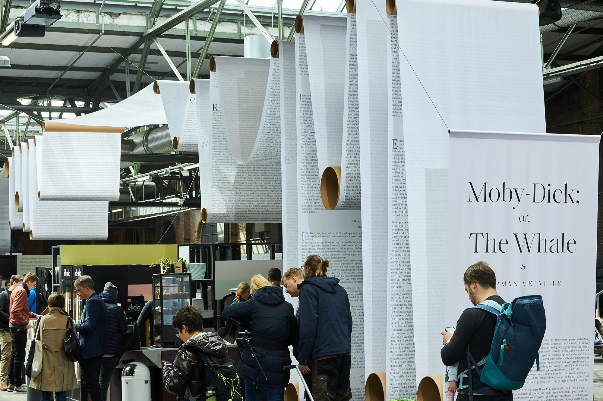 Indoor exhibition space featuring suspended paper scrolls with printed text, a large Moby Dick poster, and visitors walking through the display.