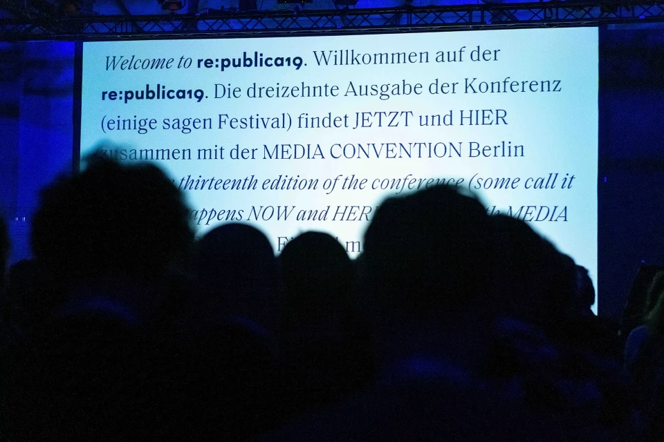 Audience at re:publica19 conference in Berlin, viewing a presentation screen with event information in German and English, bathed in blue light.