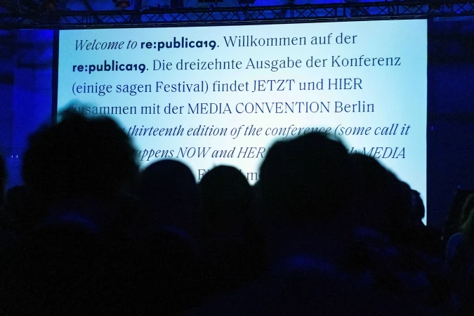 Audience at re:publica19 conference in Berlin, viewing a presentation screen with event information in German and English, bathed in blue light.