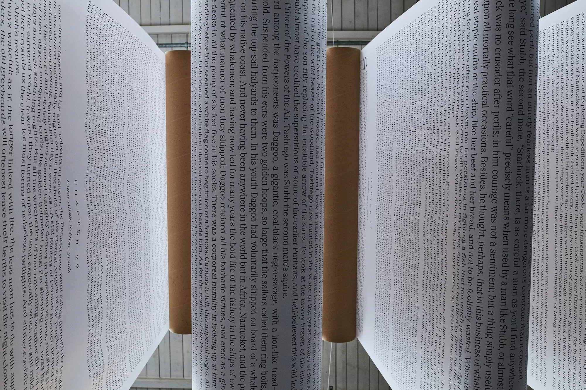 Text art installation featuring open books suspended with metal rods, displaying pages filled with dense lines of printed words.