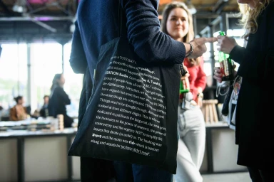 Close-up of a person wearing a blue sweater carrying a black typographic tote bag at an event.