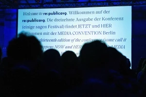 Silhouetted audience members facing a large projection screen displaying bilingual text for re-publica conference