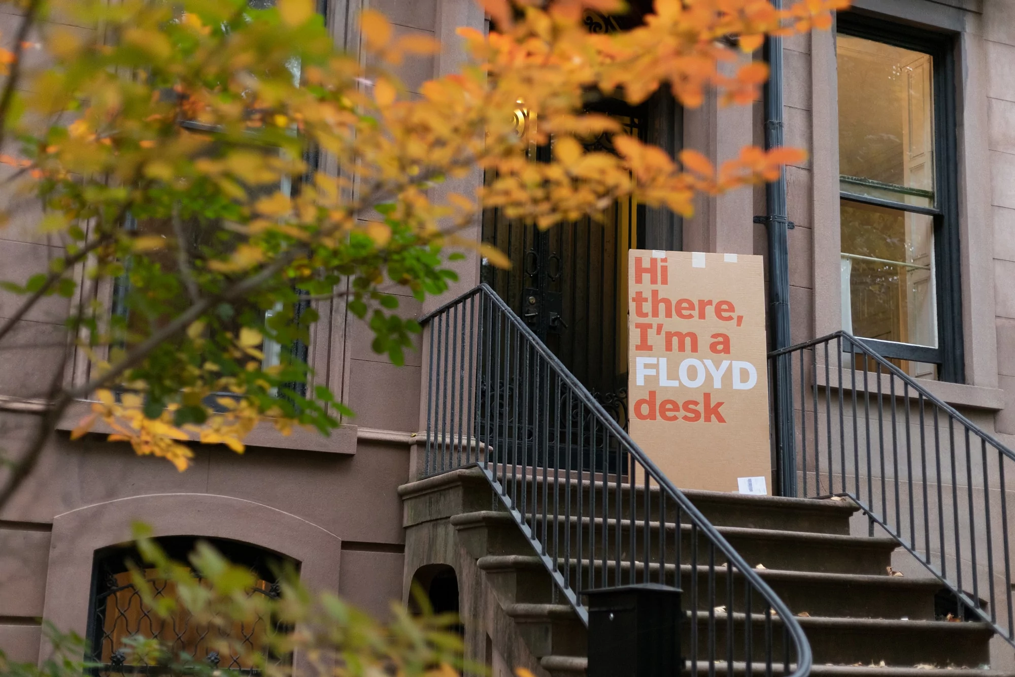 Cardboard box labeled 'Floyd Desk' sits on the steps of a brownstone in an urban setting with autumn foliage visible in the background.