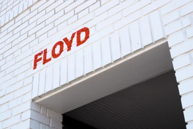 Red brand typography spelling FLOYD mounted on a white brick architectural exterior wall above a dark entryway.